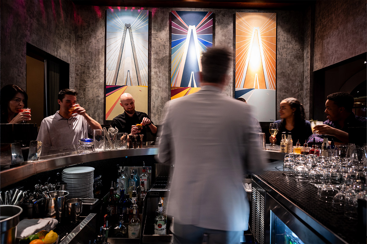 The bar at The Establishment, framed by three large, modern paintings of the Ravenel Bridge, with guests seated at the bar enjoying various cocktails and wine during happy hour on Broad Street